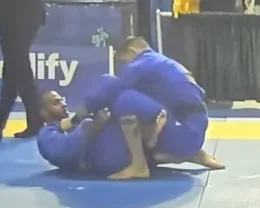 Two grapplers in blue gis at a tournament, one working from guard on the mat